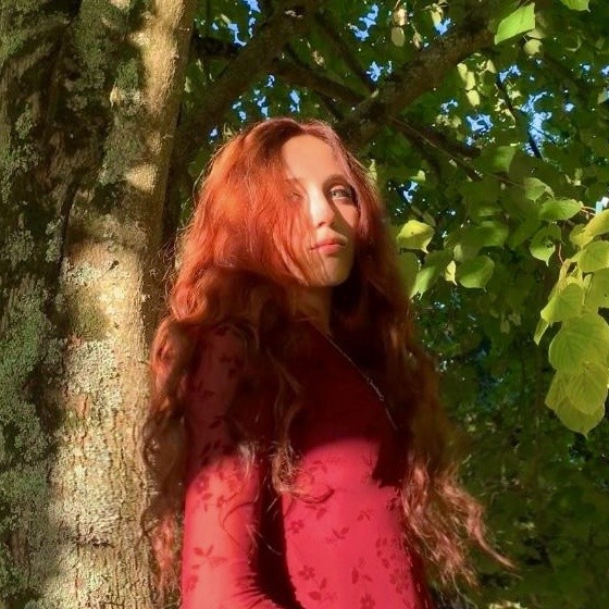 Portrait de Lola Benenati en extérieur devant un arbre, longs cheveux roux, haut rouge.